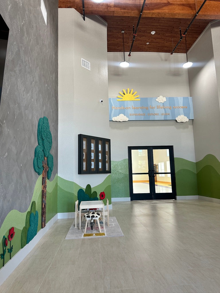Tall Montessori school lobby with yarn tree wall art, green hill murals, and a sun and cloud installation above the doors.