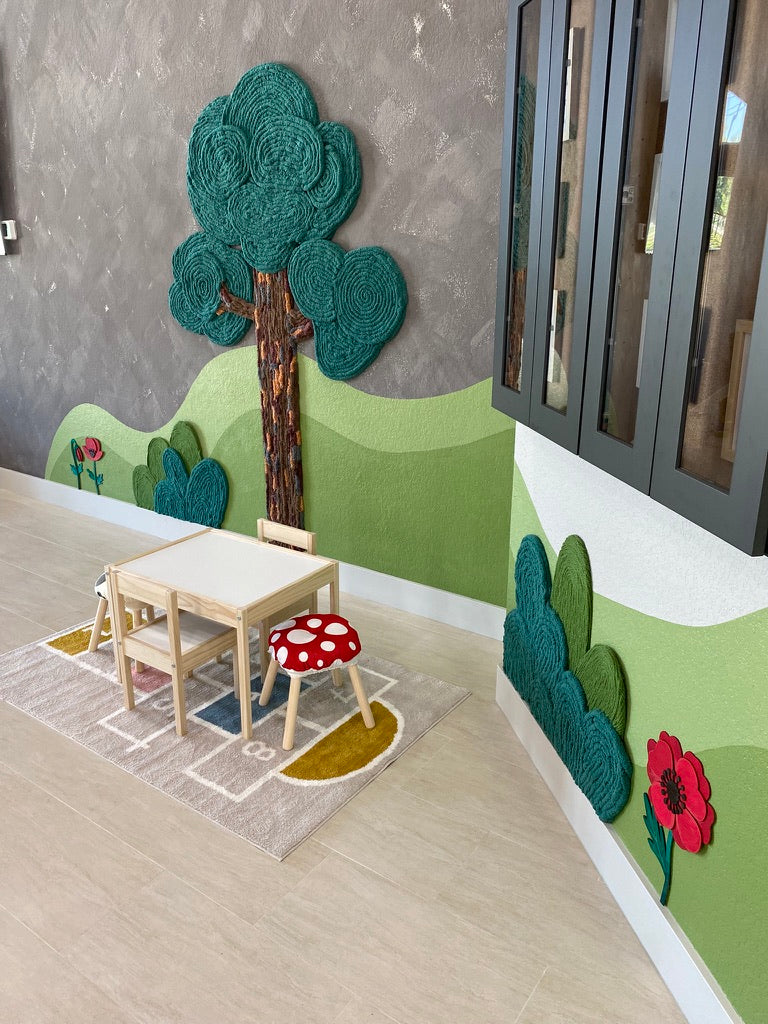 Child-sized table and mushroom stool beneath a handmade yarn tree beside red flower and shrub art in a Montessori lobby.