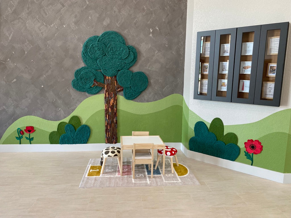 Yarn tree, red flowers, and a green hill mural surround a child-sized table in the Pinnacle Montessori school lobby.