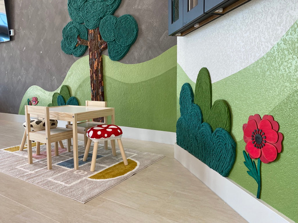 Children's table and red mushroom stool beside yarn shrubs, a red flower, and a rolling hill mural in a Montessori lobby.