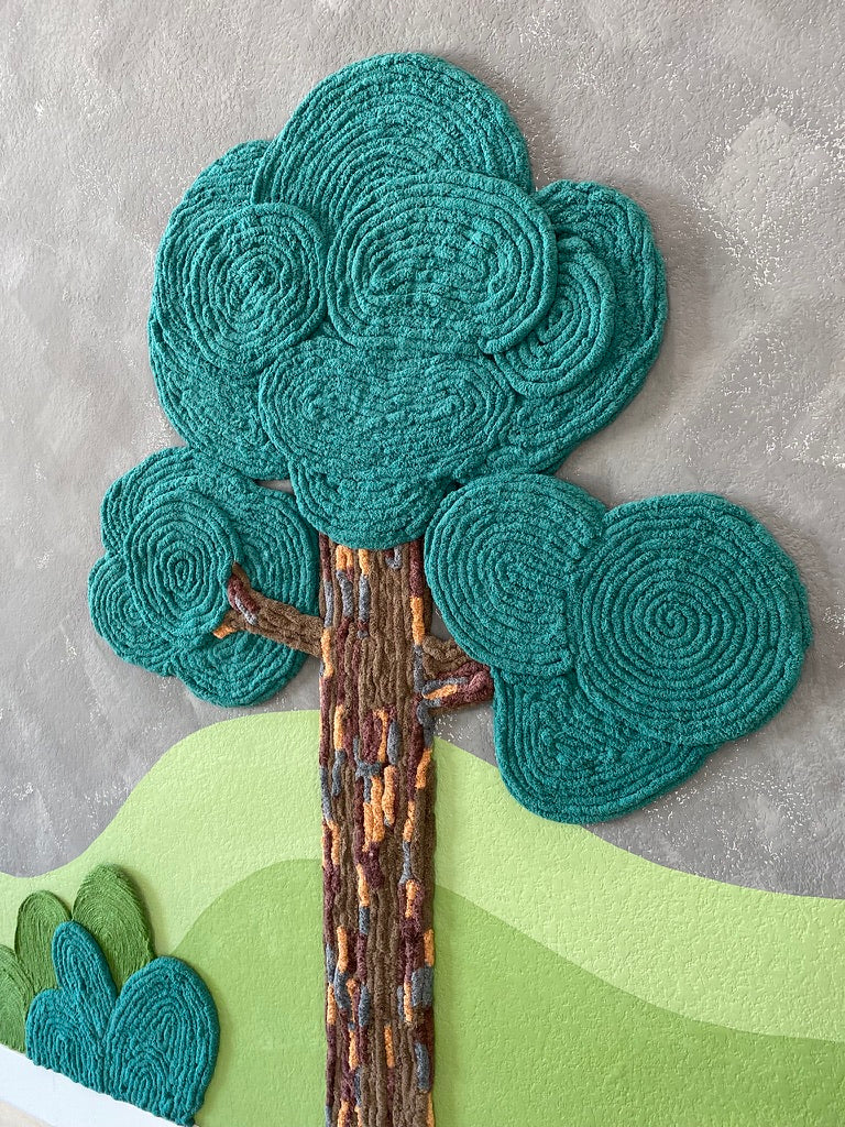 Close-up of a dimensional yarn tree with rounded teal canopies and a textured brown trunk over a green hill mural.