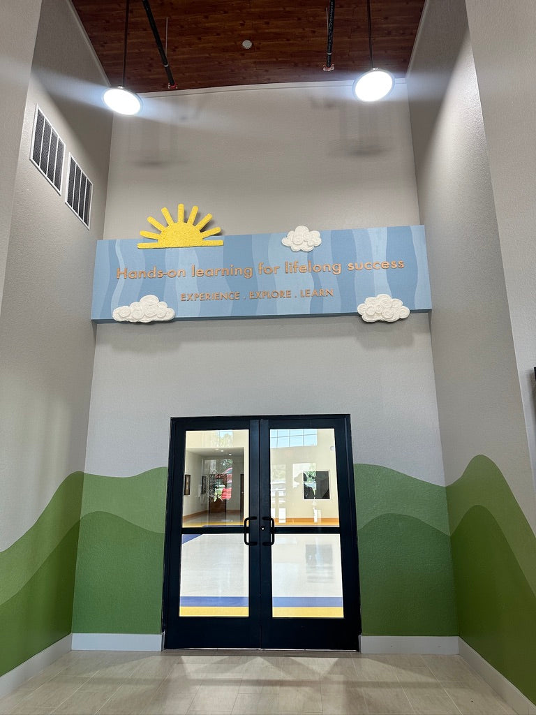 Sun and dimensional cloud artwork above the Pinnacle Montessori entrance doors, framed by a green hill mural.