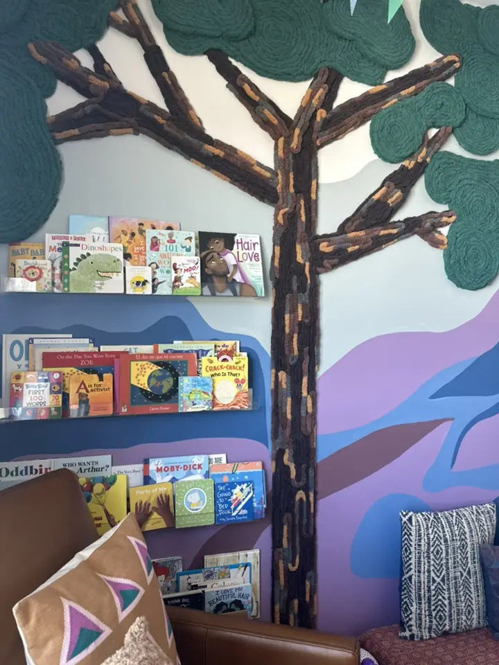 Textured yarn tree branches frame children's bookshelves and a reading chair in a colorful playroom.