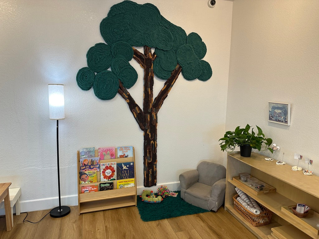 Handmade green yarn tree above bookshelves and a child-sized armchair in an Arcadia Montessori classroom reading corner.