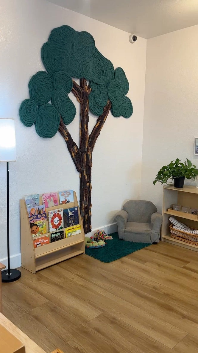 Full-height view of a textured yarn tree with spreading green canopies beside a Montessori classroom reading chair and books.