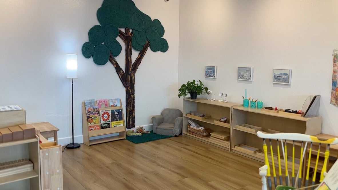 Montessori classroom with wooden learning shelves, open floor space, and handmade yarn tree art above a cozy reading corner.