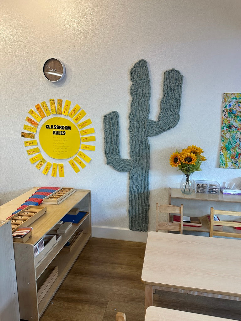 Sage-green dimensional yarn cactus beside low wooden activity shelves and a classroom rules display in a Montessori classroom.