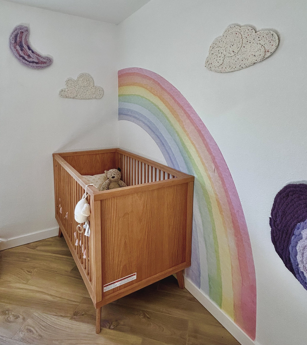 Wooden crib beneath a pastel rainbow mural, cream yarn clouds, and a lilac crescent moon in a colorful nursery.