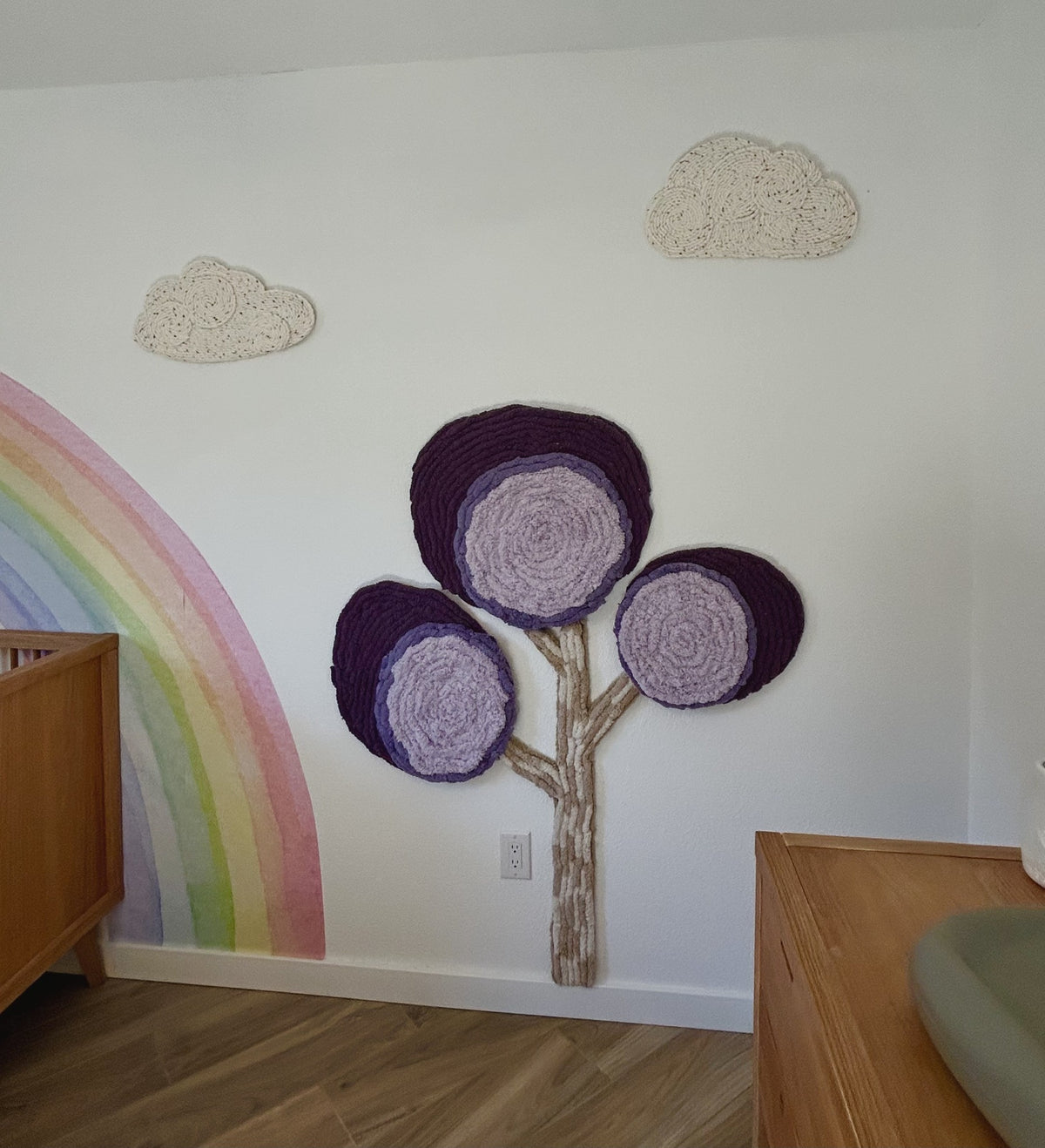 Purple and lilac dimensional yarn tree beside a pastel rainbow mural and cream clouds in a nursery.