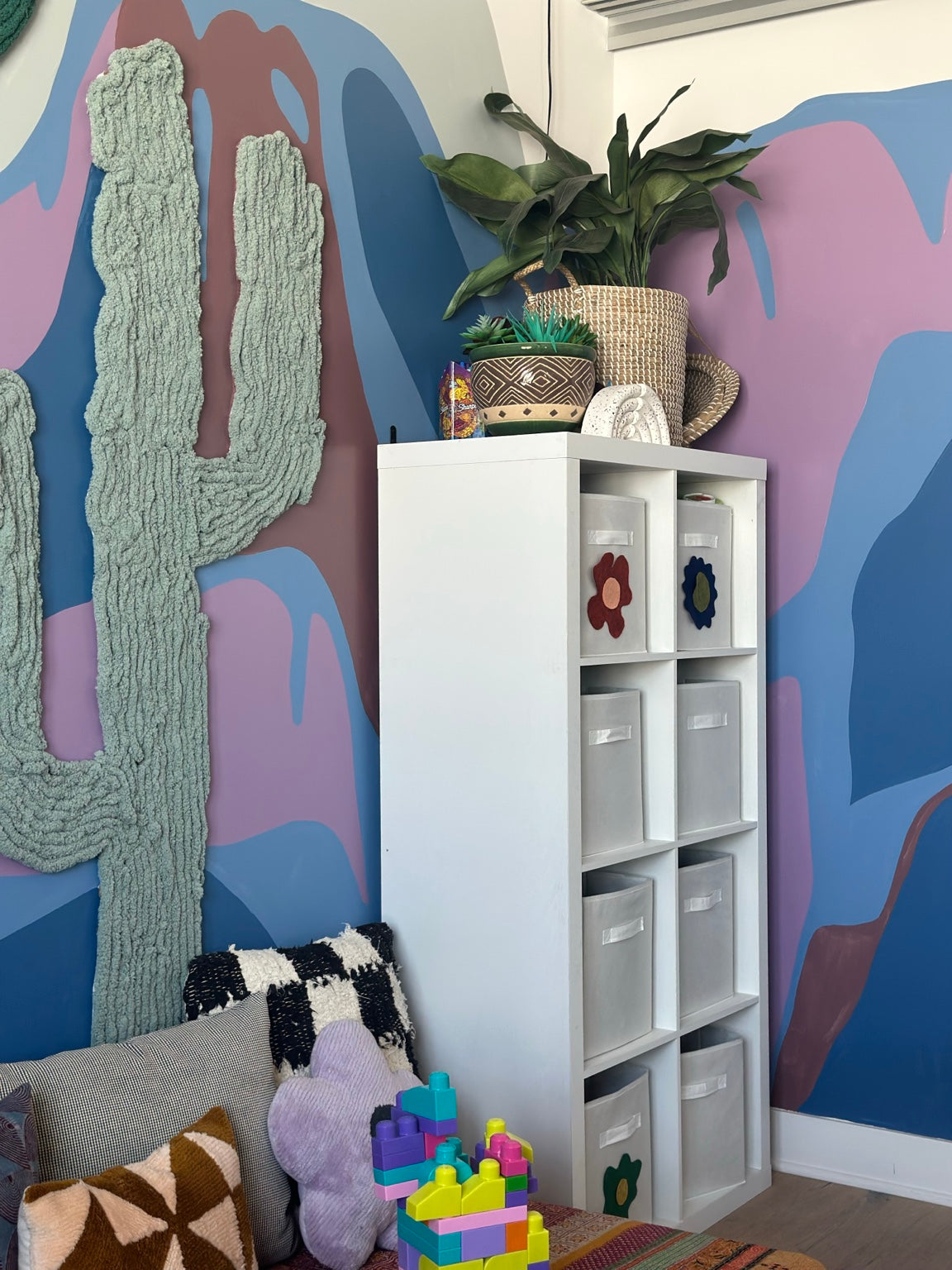 Textured yarn cactus beside plants, flower-decorated storage bins, and cushions against a colorful playroom mountain mural.