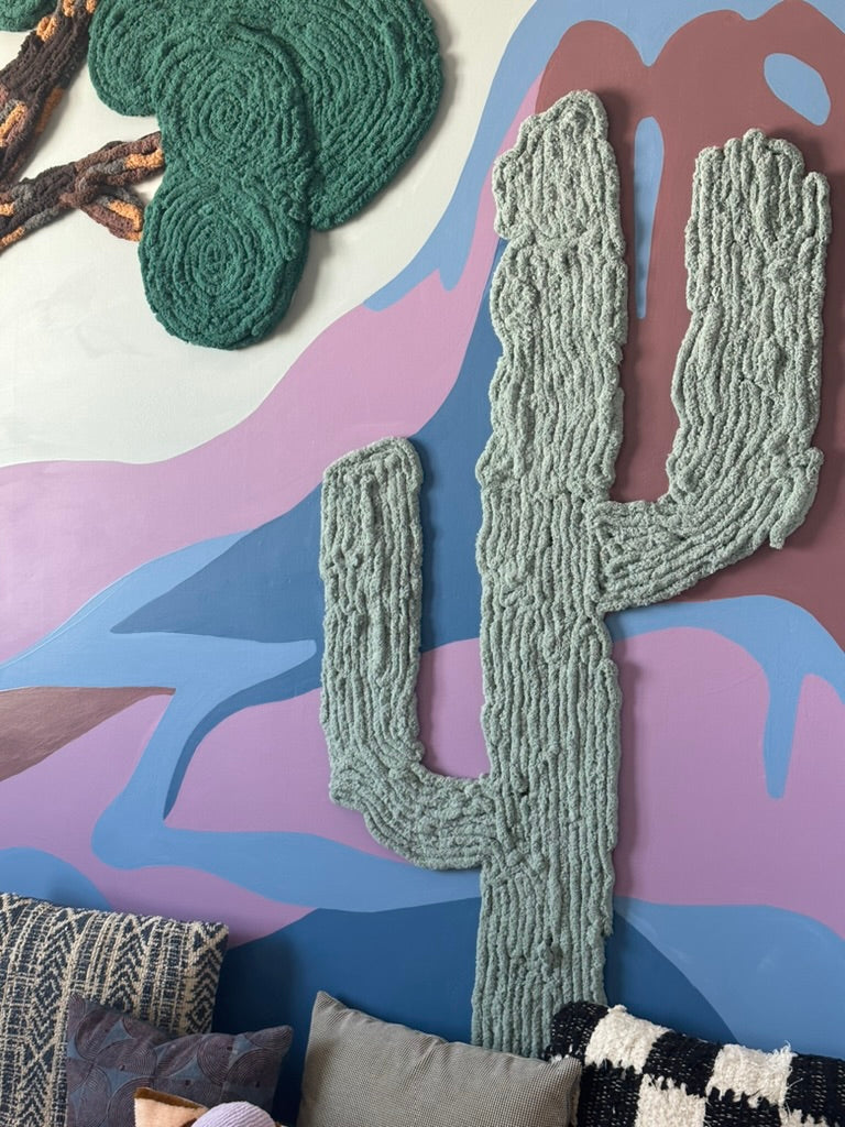 Sage-green yarn cactus and deep-green tree canopy against a pink, blue, and mauve mountain mural above playroom cushions.