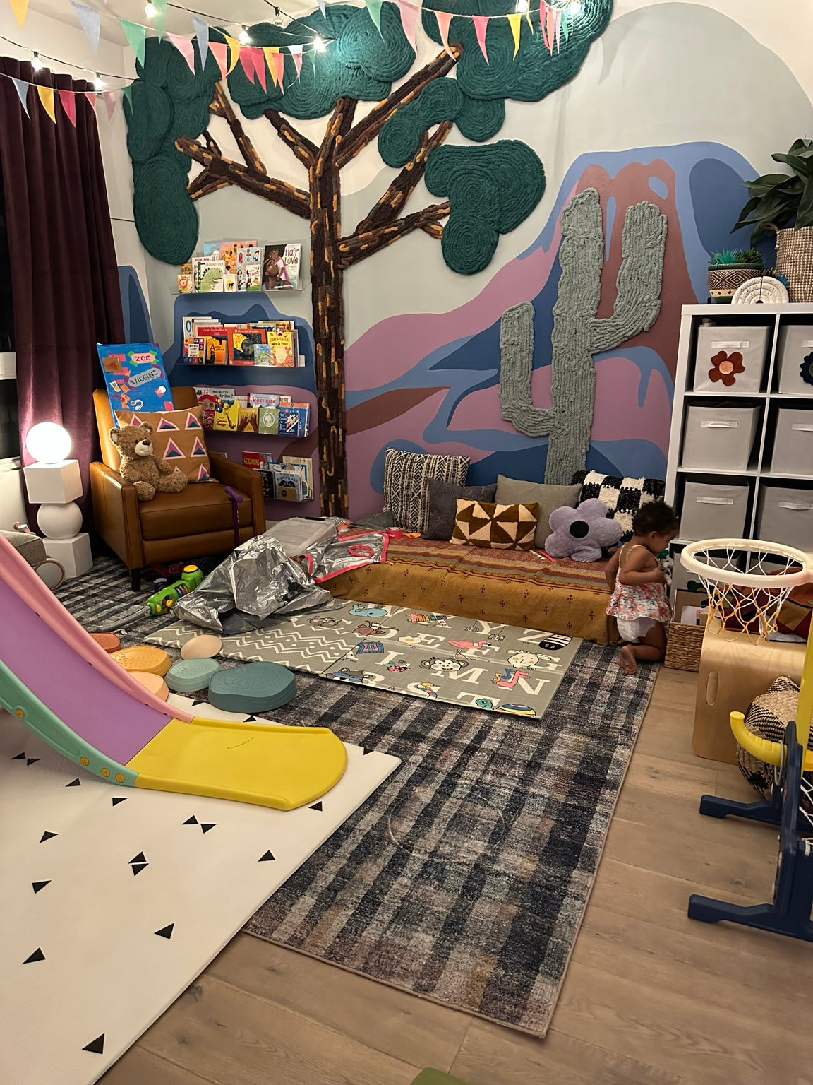 Family playroom with a rainbow slide, reading nook, and dimensional yarn tree and cactus over a colorful mountain mural.