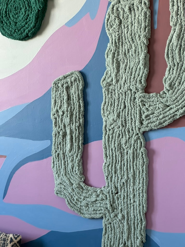 Close-up of soft sage-green yarn cactus arms against a layered pink, blue, and mauve mountain mural.