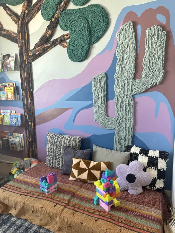 Handmade yarn tree and sage-green cactus above a playroom reading bench, with a colorful mountain mural and cozy cushions.