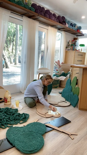 Meg applying adhesive to a wooden tree shape, surrounded by green coiled-yarn canopies and handmade shrubs in her studio.