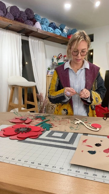 Meg assembling painted flower wall art at a worktable beneath shelves of colorful yarn in the That Playful Life studio.