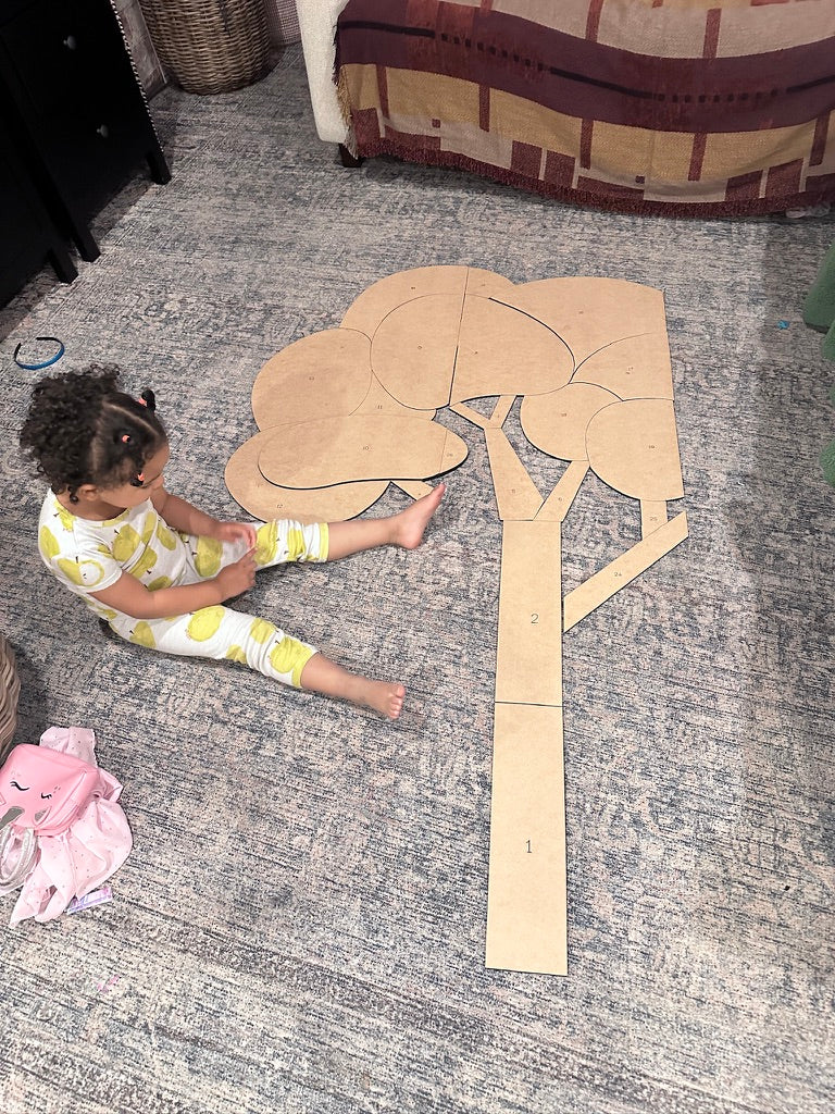 A child sitting beside numbered wooden trunk, branch, and canopy pieces laid out for a handmade sensory tree installation.
