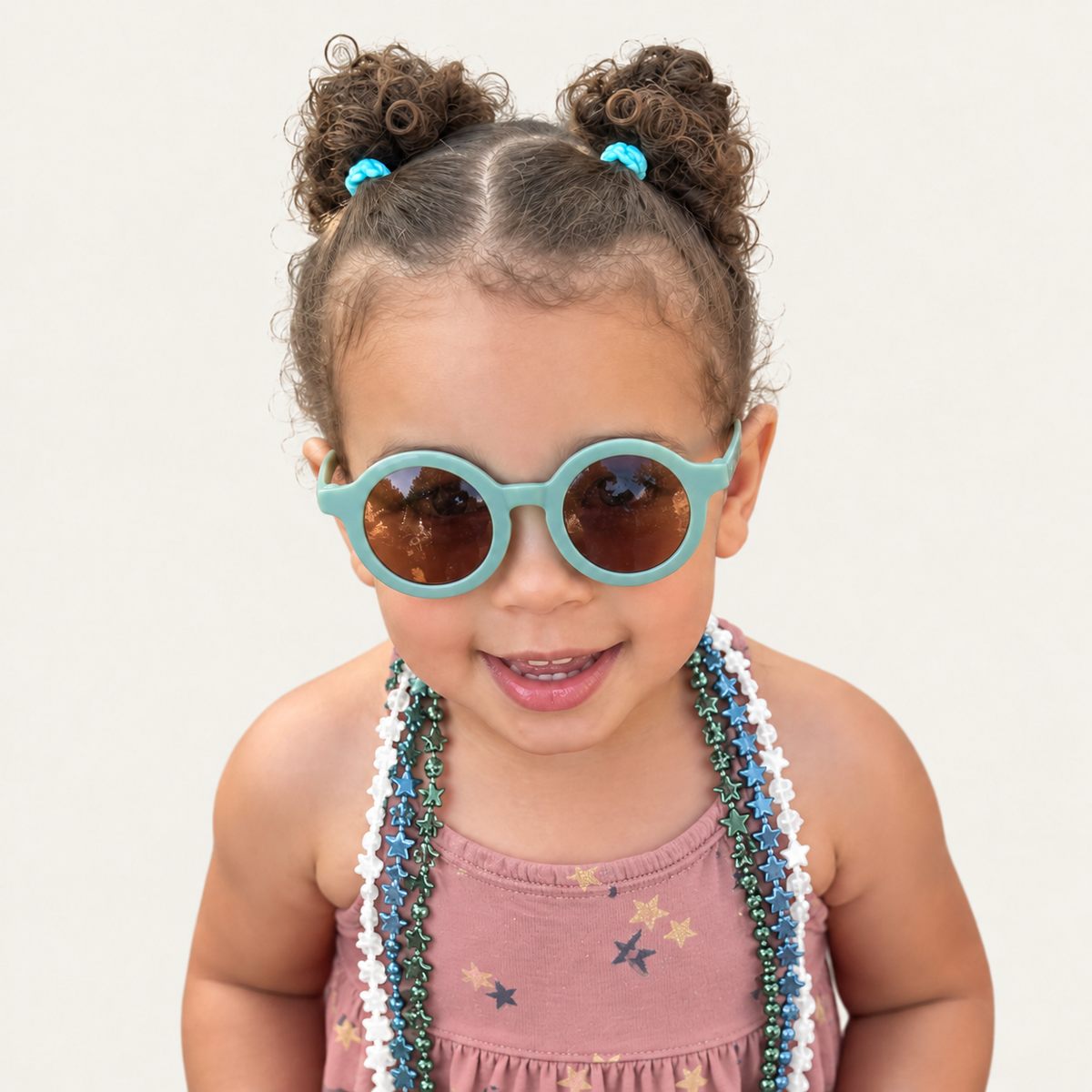 Rae smiling in turquoise sunglasses and colorful bead necklaces against a beige background.