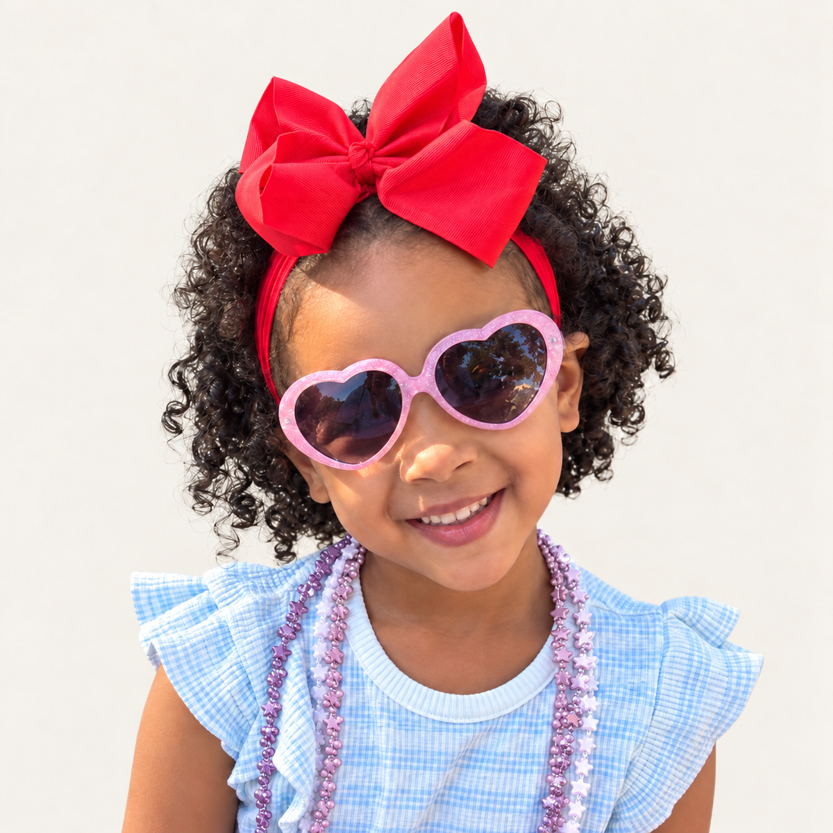 Zoe smiling in heart-shaped sunglasses and a red bow against a beige background.